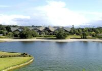 Vista da lagoa, faixa de areia e o clube principal do Condomínio Bosques de Atlântida, com telhados escuros e arquitetura ...