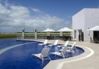 Piscina azul com espreguiçadeiras submersas e deck no Condomínio Costa Serena. Edifício moderno e paisagem natural.