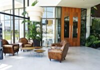 Lobby com grandes janelas de vidro, poltronas de couro marrom, mesas de centro redondas, plantas verdes e uma máquina de v...