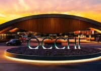 Pórtico de entrada moderno do Occhi Marina Club - Capão Novo, com letras iluminadas e paisagismo noturno, sob um céu alara...