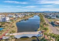 Visão panorâmica do condomínio Quintas do Lago - Xangri-Lá, exibindo lago azul, ponte branca, casas contemporâneas e palme...