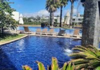 Cenário de lazer com piscina azul, cascata de pedra, espreguiçadeiras e palmeiras no Condomínio Rossi Atlântida, sob céu c...