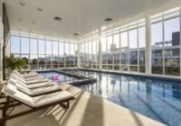 Piscina interna do Condomínio Rossi Atlântida com paredes de vidro, espreguiçadeiras e luz natural abundante, oferecendo l...